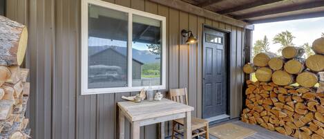 A wooden deck with a table and chair, a window, a door, and a stack of firewood.