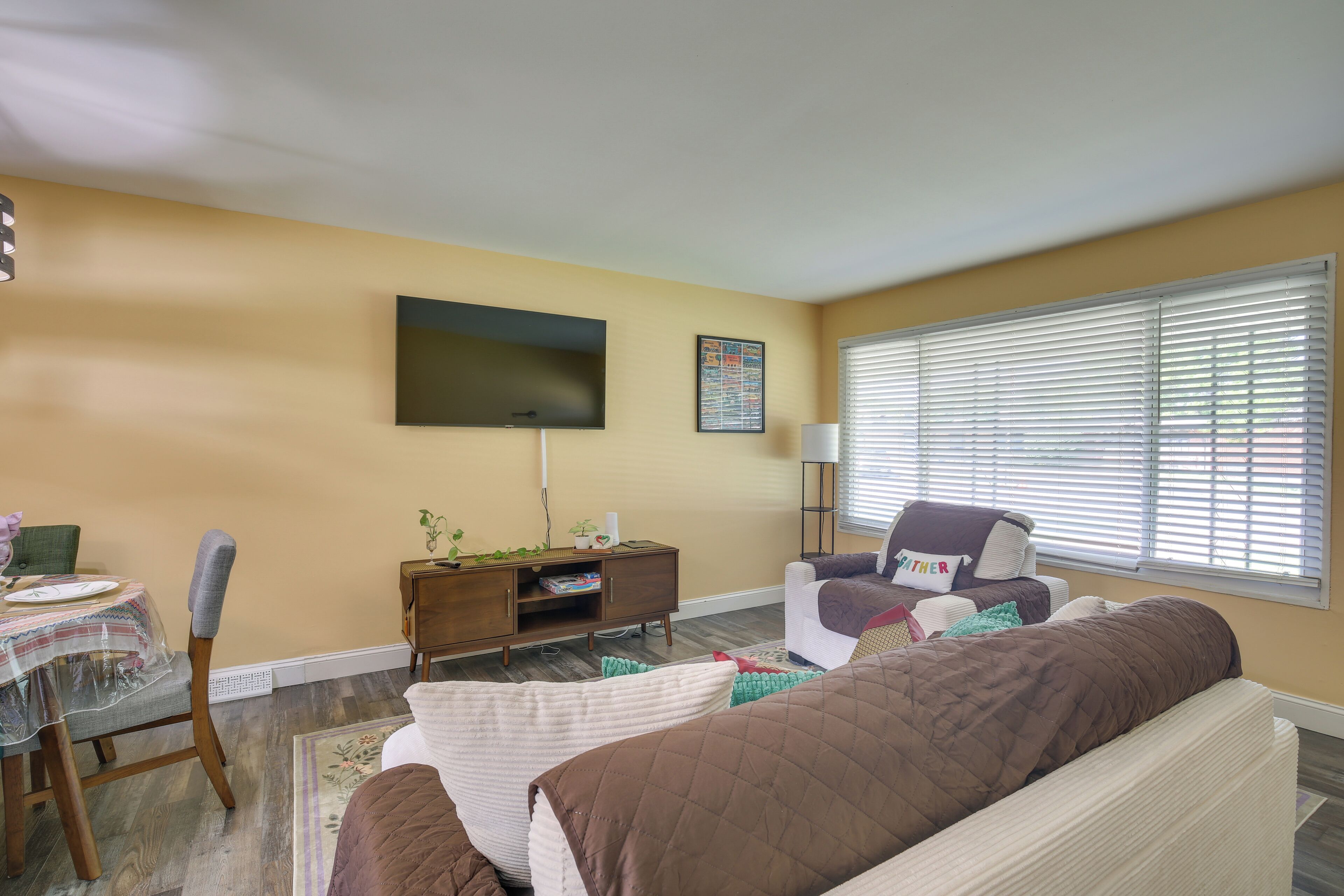 Una sala de estar con un sofá, un televisor montado en la pared, una mesita con una silla y una ventana con persianas.