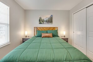 A bedroom with a green bedspread, a wooden headboard, two bedside lamps, a nightstand, and a mountain landscape painting on the wall. - Home Near Air Force, Great Wolf, Ford Amphitheater (Colorado Springs)
