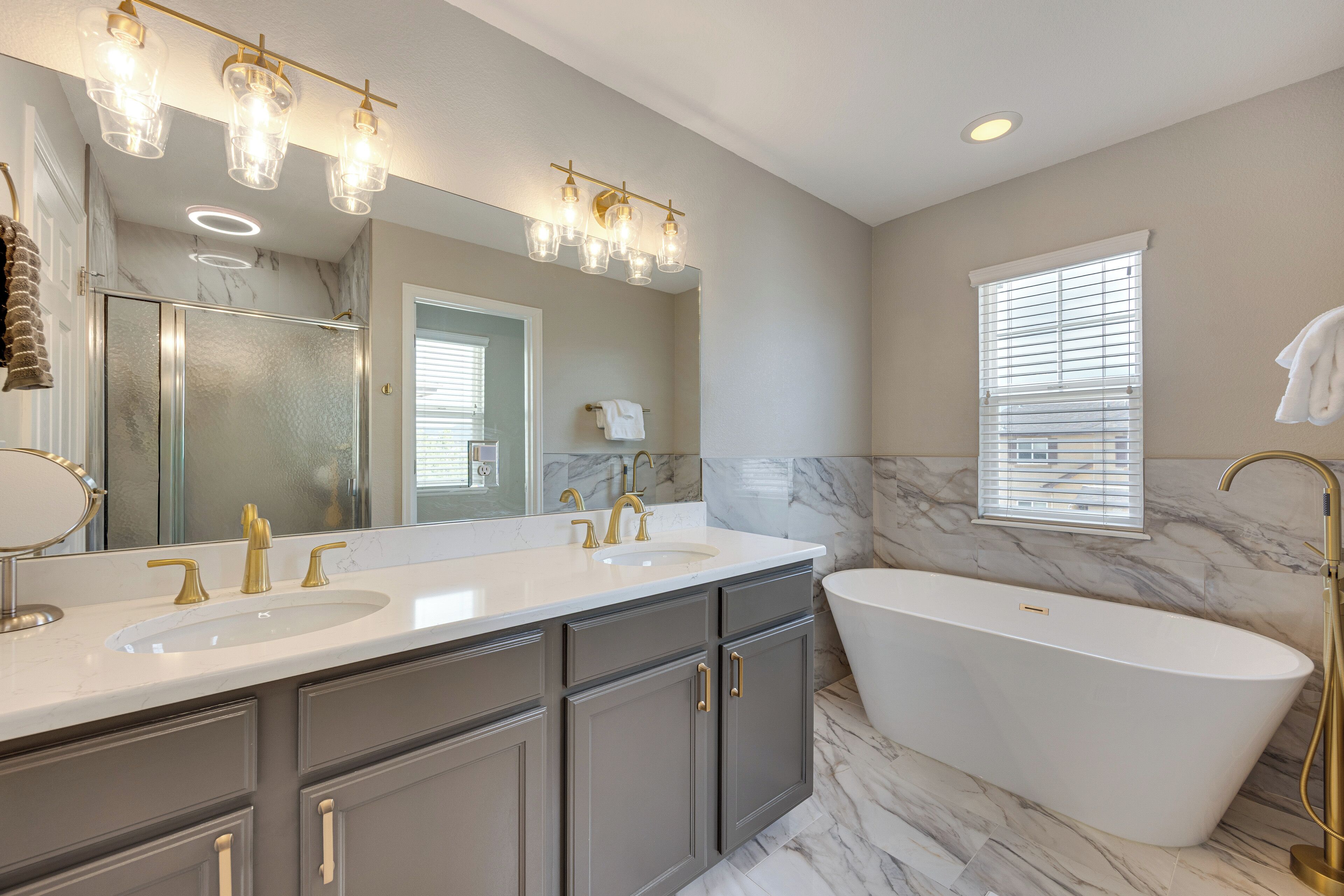 A bathroom with a double sink vanity, a freestanding bathtub, a large mirror, and a window with blinds.