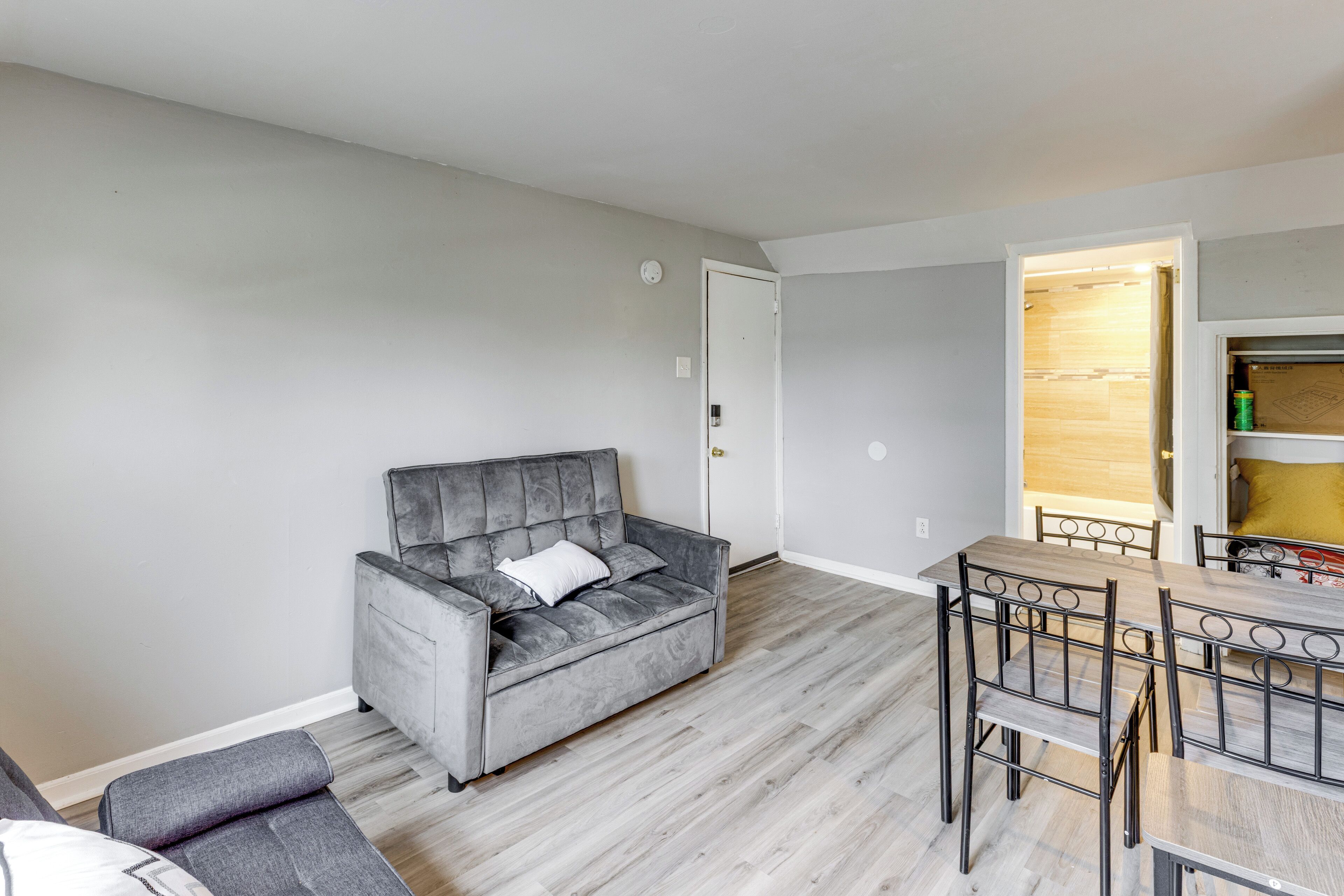Una sala de estar con un sofá gris, una mesa de comedor con sillas, y un área de cocina visible a través de una puerta abierta.