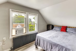 A bedroom with a bed, two windows, a radiator, and a view of greenery outside.