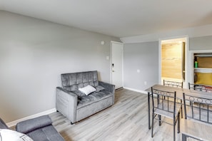 A living room with a grey sofa, a dining table with chairs, and a kitchen area visible through an open door.