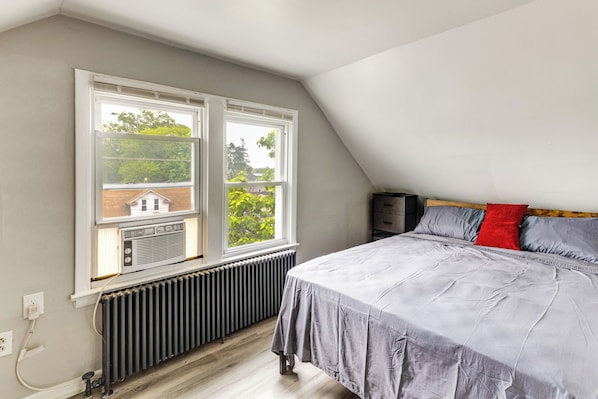 A bedroom with a bed, two windows, a radiator, and a view of greenery outside.