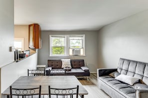 A modern living room with a sofa, a dining table, and chairs. There are two windows with air conditioning units.