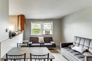 Uma sala de estar moderna com um sofá, uma mesa de jantar e cadeiras. Há duas janelas com aparelhos de ar condicionado.