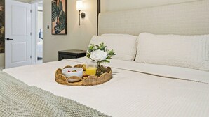 Un lit bien fait, recouvert d’une couette blanche, un plateau en osier contenant des éléments de petit-déjeuner et un luminaire fixé au mur.