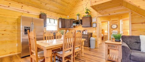 A wooden interior with a dining table, chairs, a refrigerator, a microwave, cabinets, a clock, and a sofa.