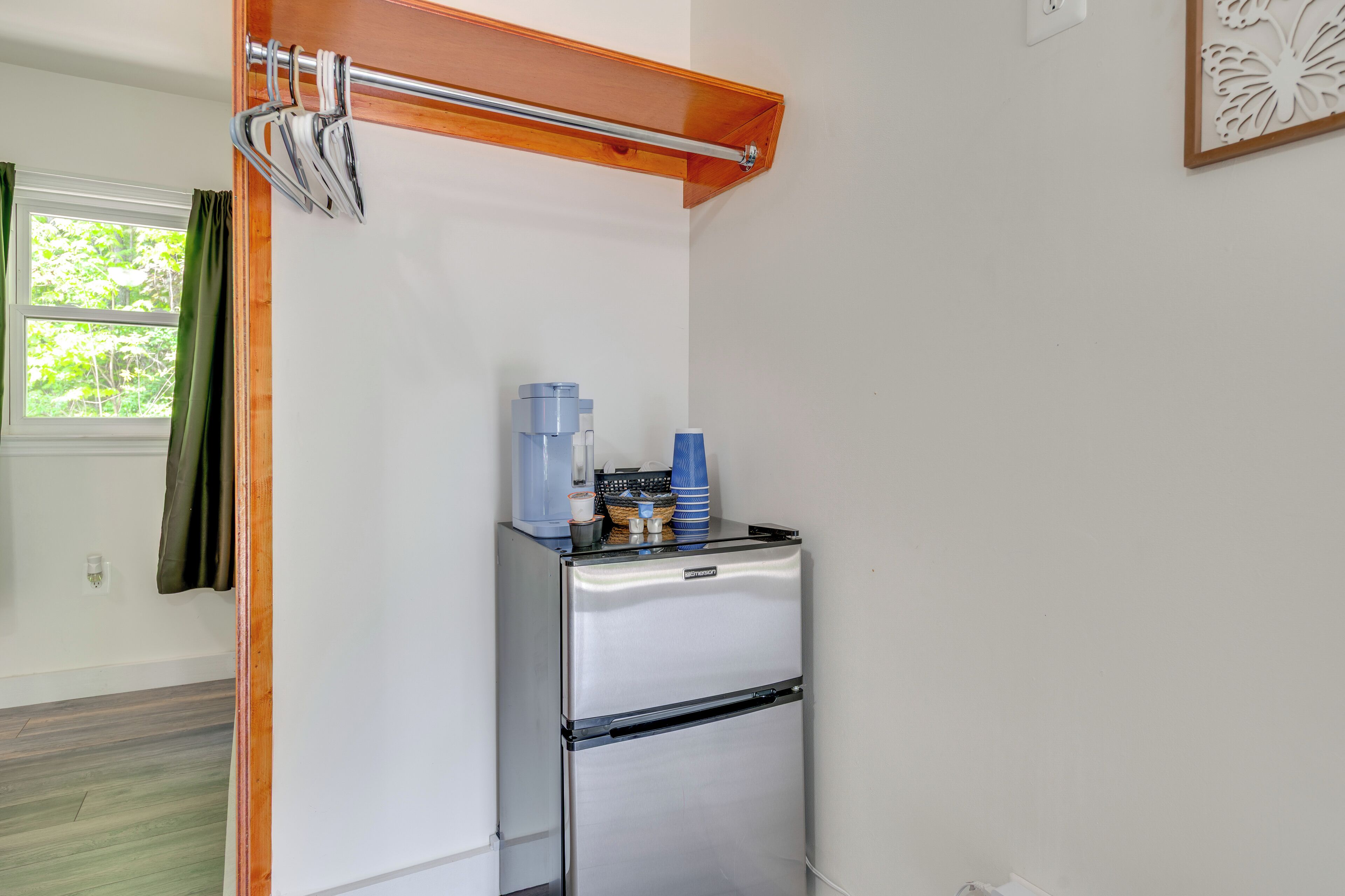 Una cocina compacta con un refrigerador de acero inoxidable, un estante de madera encima y una ventana con cortinas.