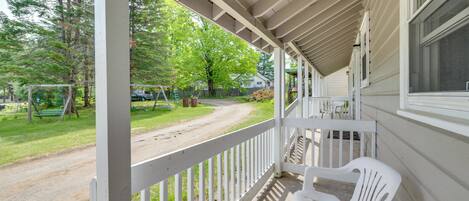 A covered porch with a white railing, a white plastic chair, and a view of a park with a swing set and trees.