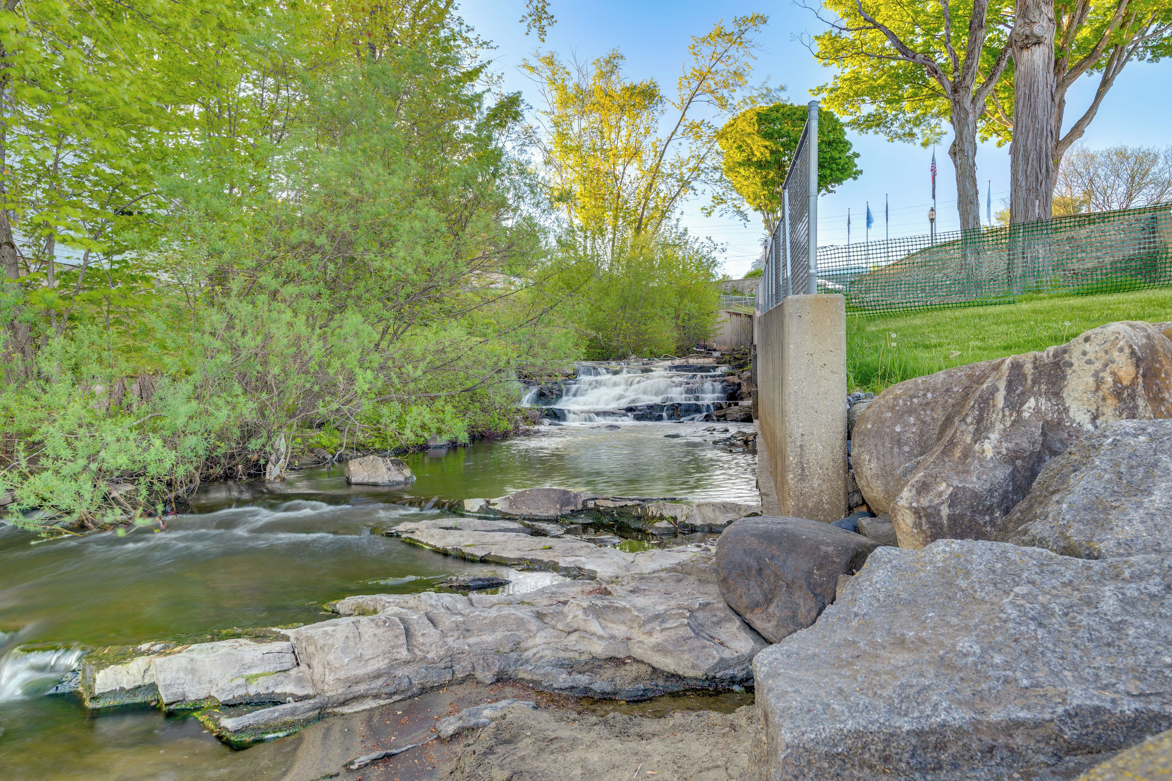 A natural stream with rocks and trees, a concrete structure, and a fence in the background.