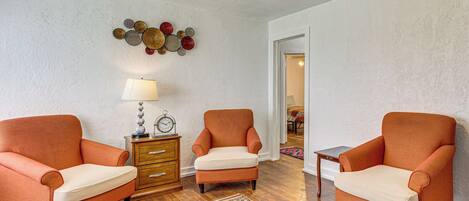Quarto com duas poltronas laranja, uma mesa de apoio de madeira com abajur, tapete estampado e ventoinha no teto.