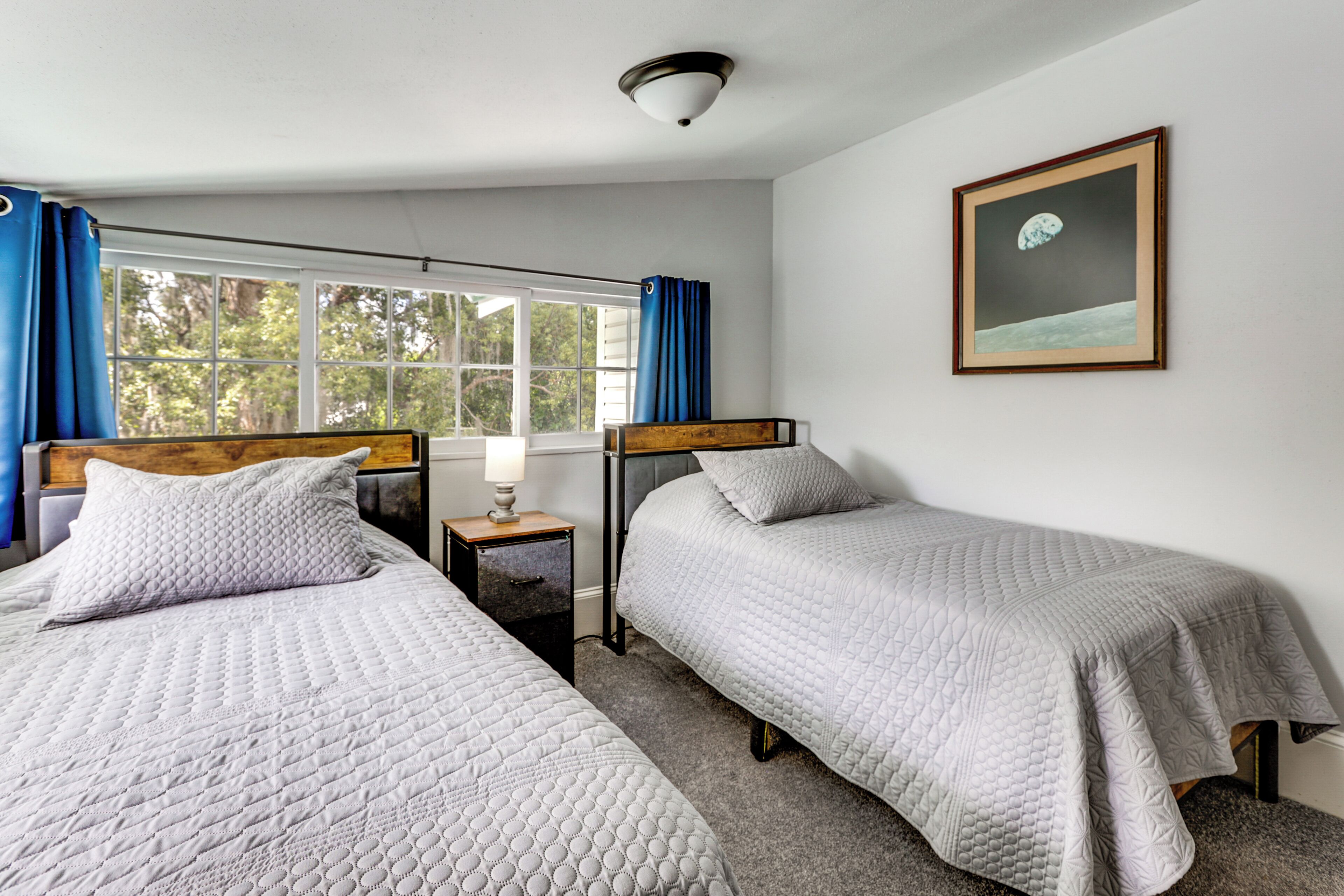A room with two beds, a nightstand with a lamp, a framed picture on the wall, and a window with curtains.