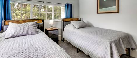 A room with two beds, a nightstand with a lamp, a framed picture on the wall, and a window with curtains.