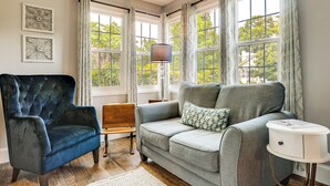 A room with a dark blue armchair, a grey sofa with patterned pillows, a small wooden bench, and a white side table with a lamp. - Walk to Stetson and Historic Downtown Deland! (DeLand)