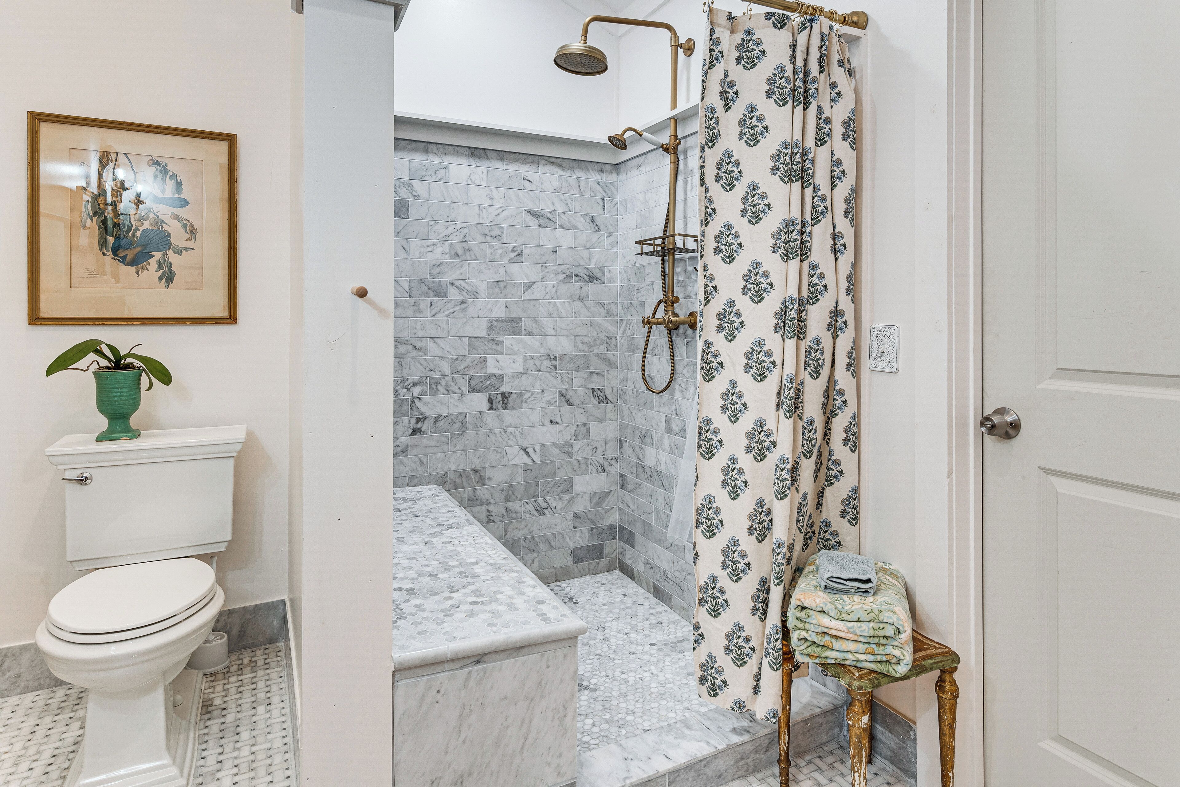 A bathroom with a toilet, shower, marble tiles, a framed artwork, and a potted plant.