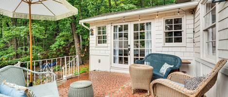 A covered patio with wicker furniture, a white umbrella, and a house with large windows.