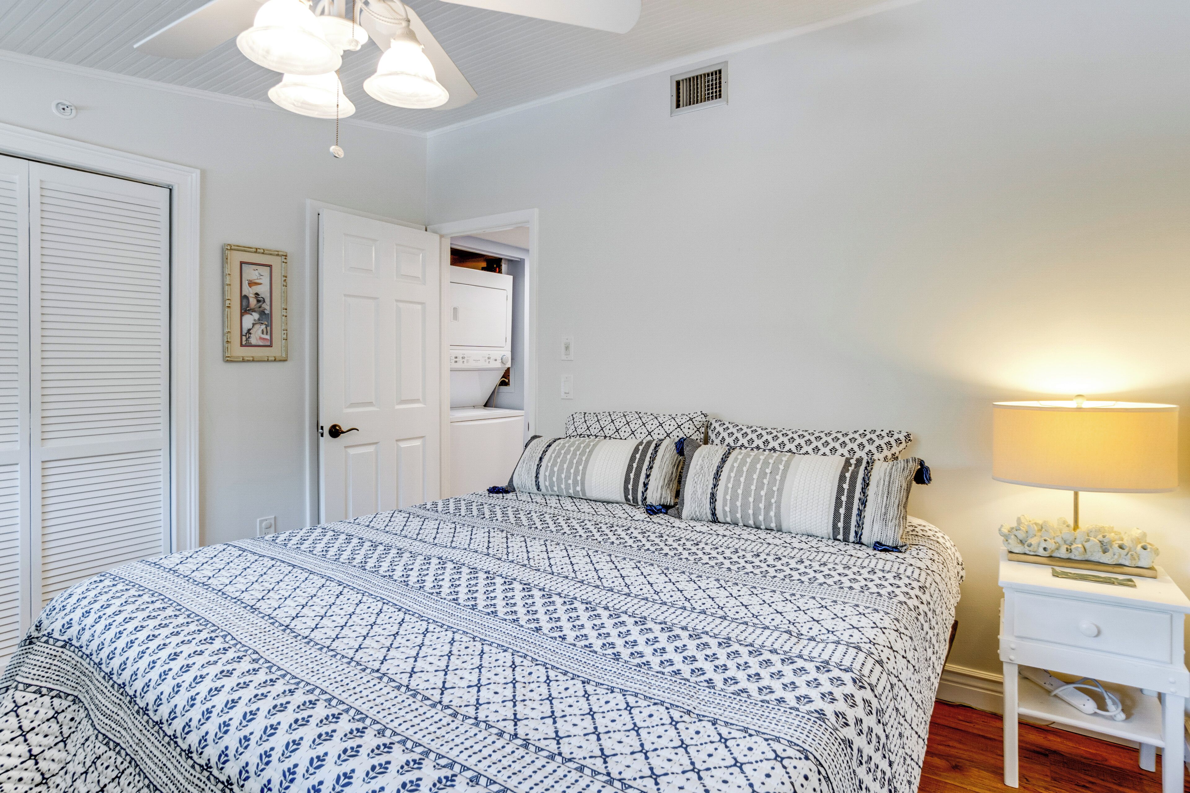 A bedroom with a bed, a nightstand, a lamp, a wardrobe, and a door leading to another room.