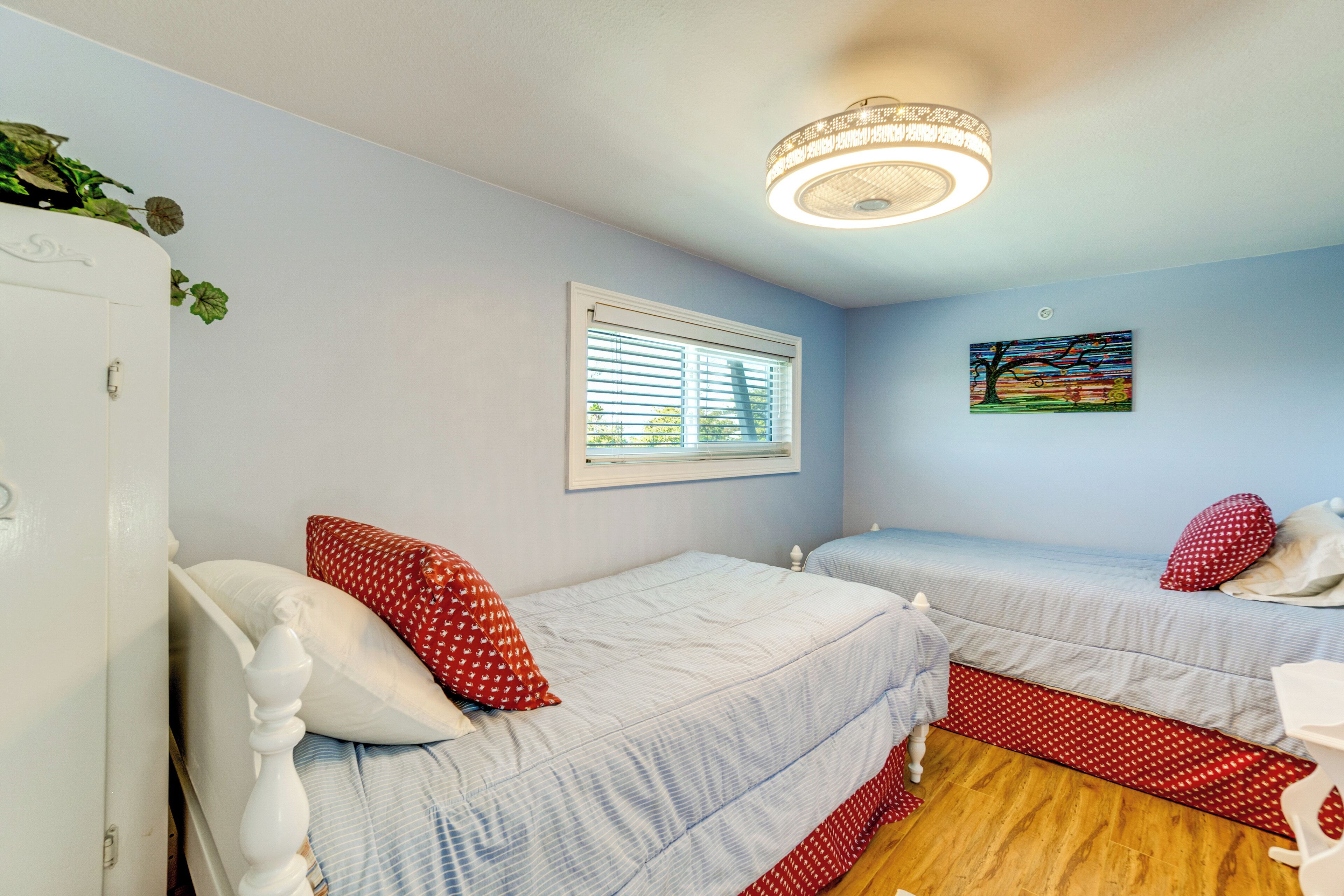 A bedroom with two beds, a window with blinds, a painting on the wall, and a ceiling light fixture.