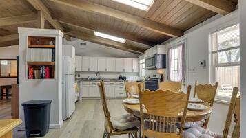 A kitchen with wooden beams, white cabinets, a dining table with chairs, and a trash bin.