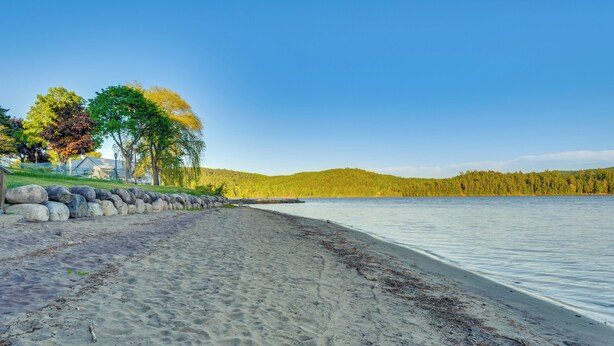 A sandy beach with a calm lake, trees, and a distant hill.