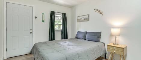 Un dormitori amb un llit, una taula de nit amb una làmpada, una porta i una finestra amb cortines.