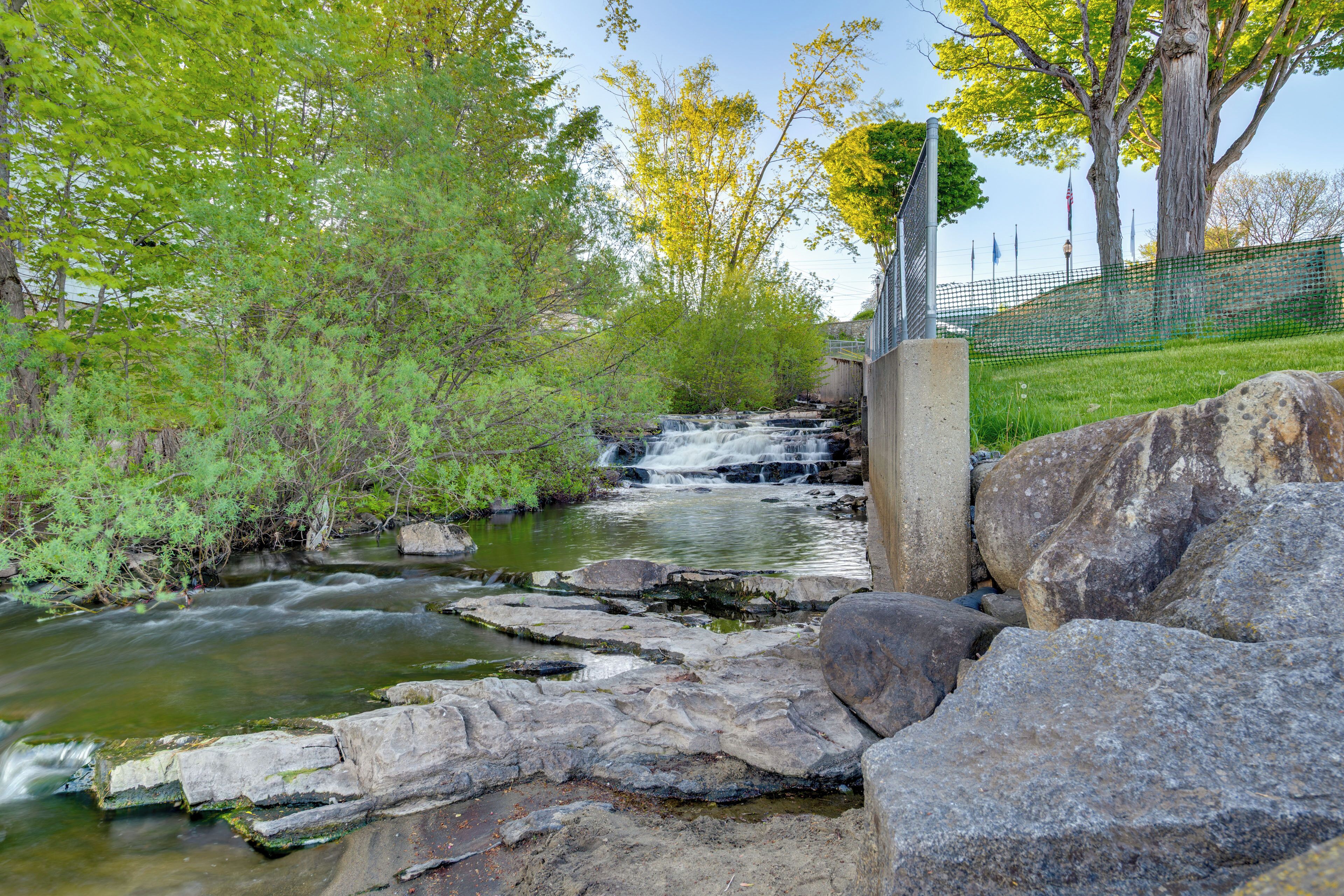 A natural stream with rocks and trees, a wooden bridge, and a fence with flags.