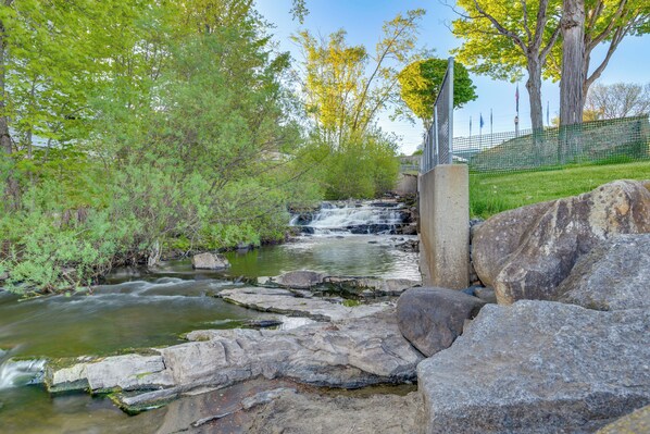 A natural stream with rocks and trees, a small waterfall, and a fence in the background.