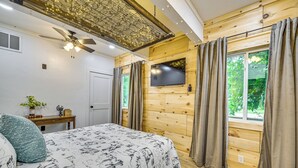 Ett sovrum med en säng, en takfläkt, en tv, träpanel på väggarna och ett fönster med gardiner.
