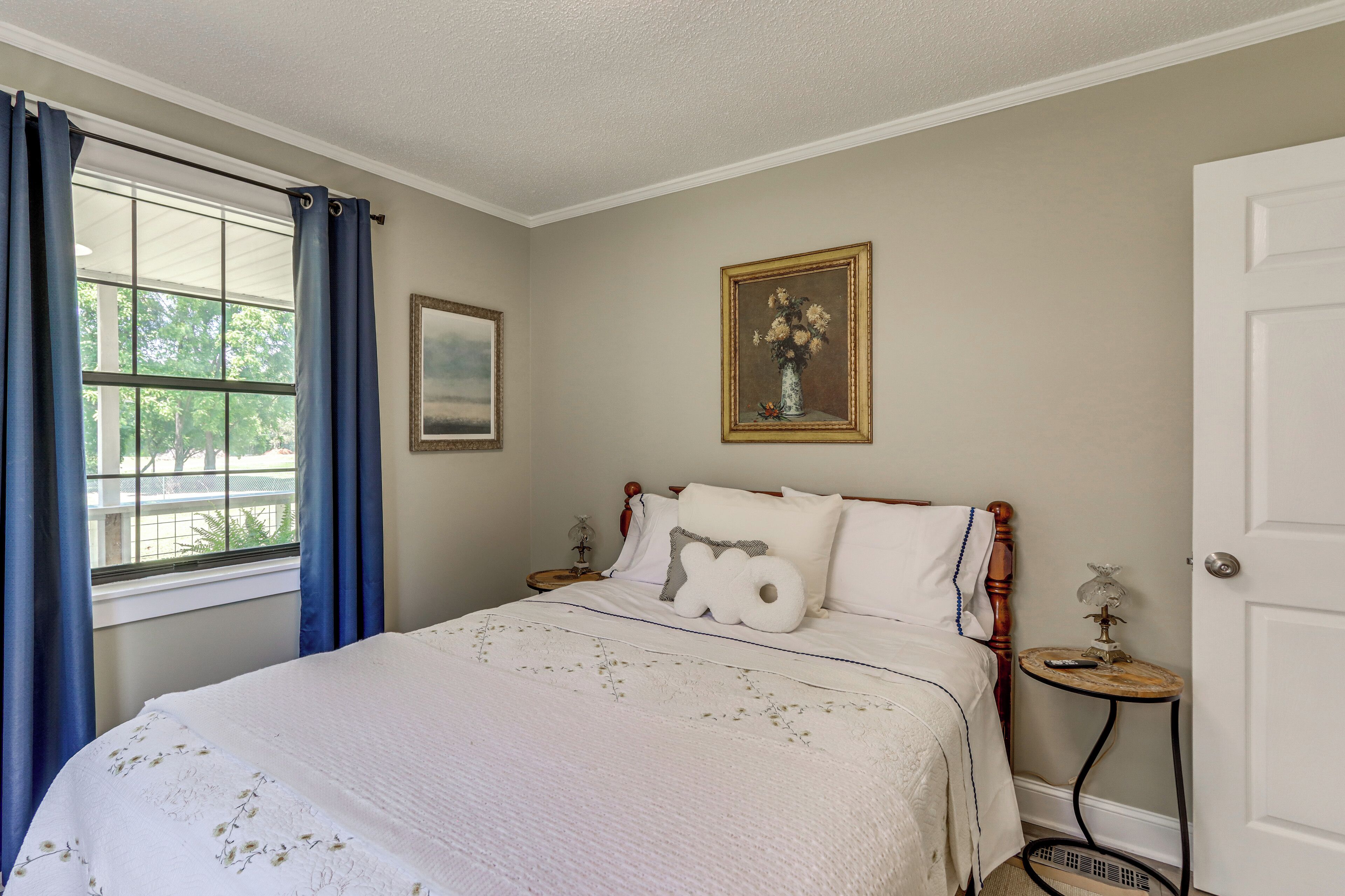 A bedroom with a bed, a nightstand, a window with curtains, a door, and a framed picture on the wall.