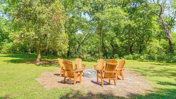 A circular seating area with chairs around a central fire pit, surrounded by a grassy field and dense trees.