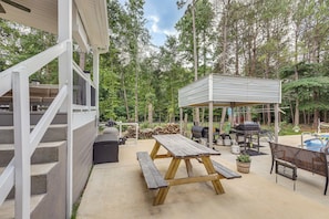 A covered outdoor patio with a wooden table and benches, a barbecue grill, and a pool.