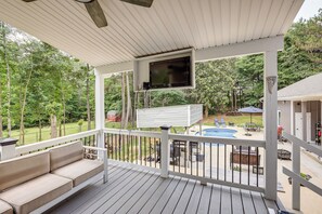 Eine überdachte Terrasse mit Fernseher, Sitzgelegenheiten im Freien und Blick auf einen Garten mit Pool und Gartenmöbeln.