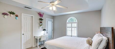 A bedroom with a bed, a desk, a chair, a ceiling fan, and a window.