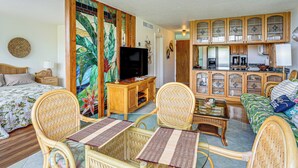 A living room with wicker furniture, a television, and a kitchen area with wooden cabinets. - Pool Access, 1 Mi to Makaha Beach! Ocean-view Unit (Waianae)