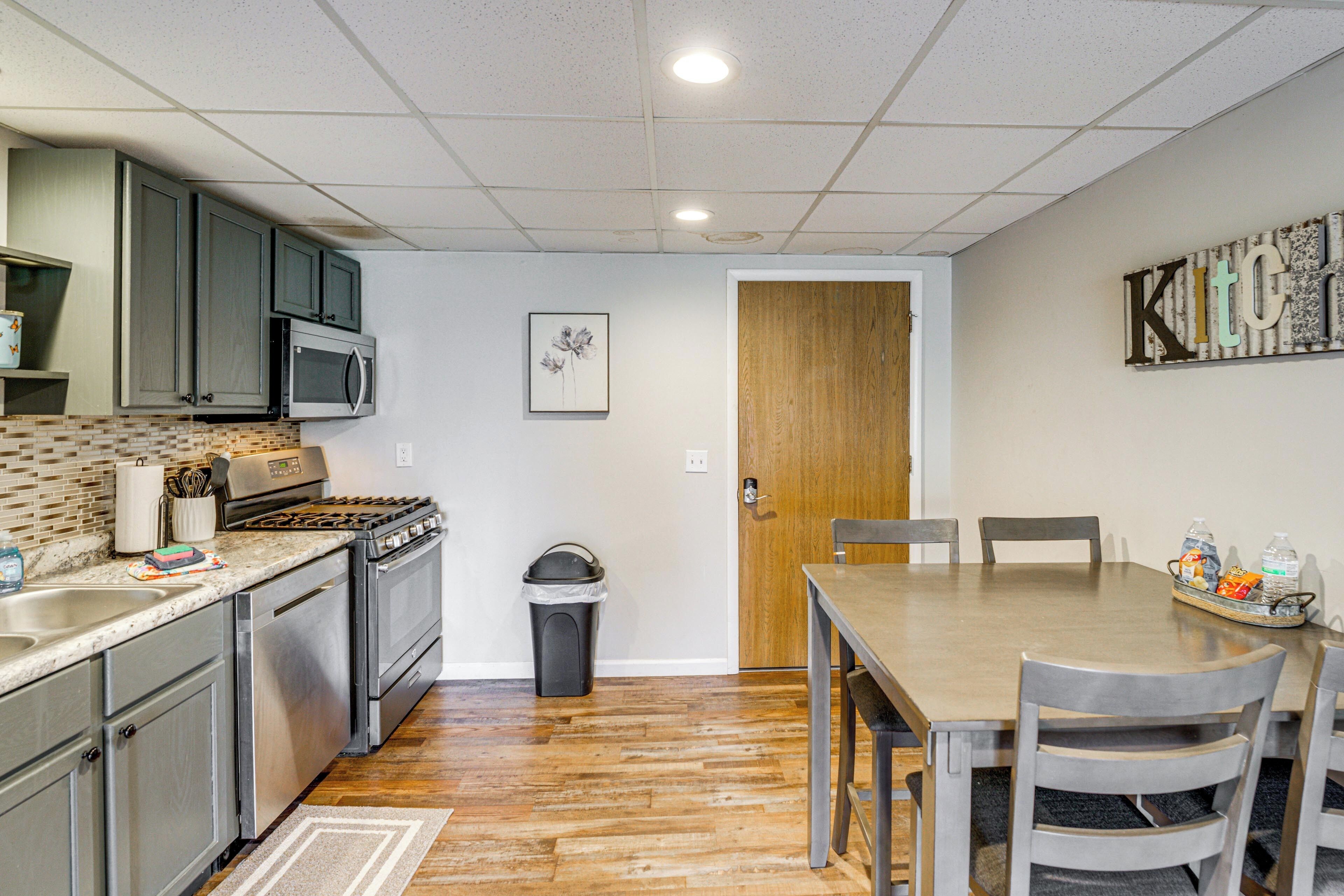 A compact kitchen with a microwave, stove, and sink. There's a dining table with chairs and a trash can.