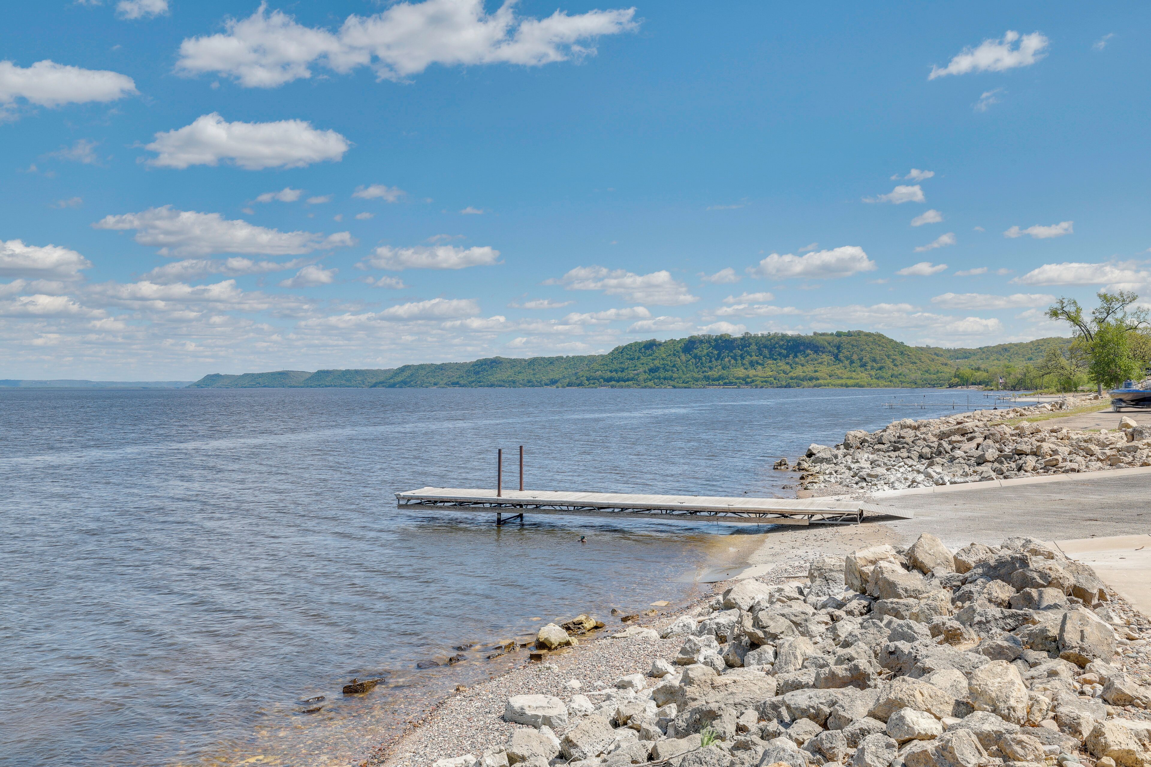 A calm lake with a dock extending into the water, surrounded by rocky terrain and a distant green hill.