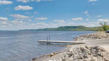 A calm lake with a dock extending into the water, surrounded by rocky terrain and a distant green hill.
