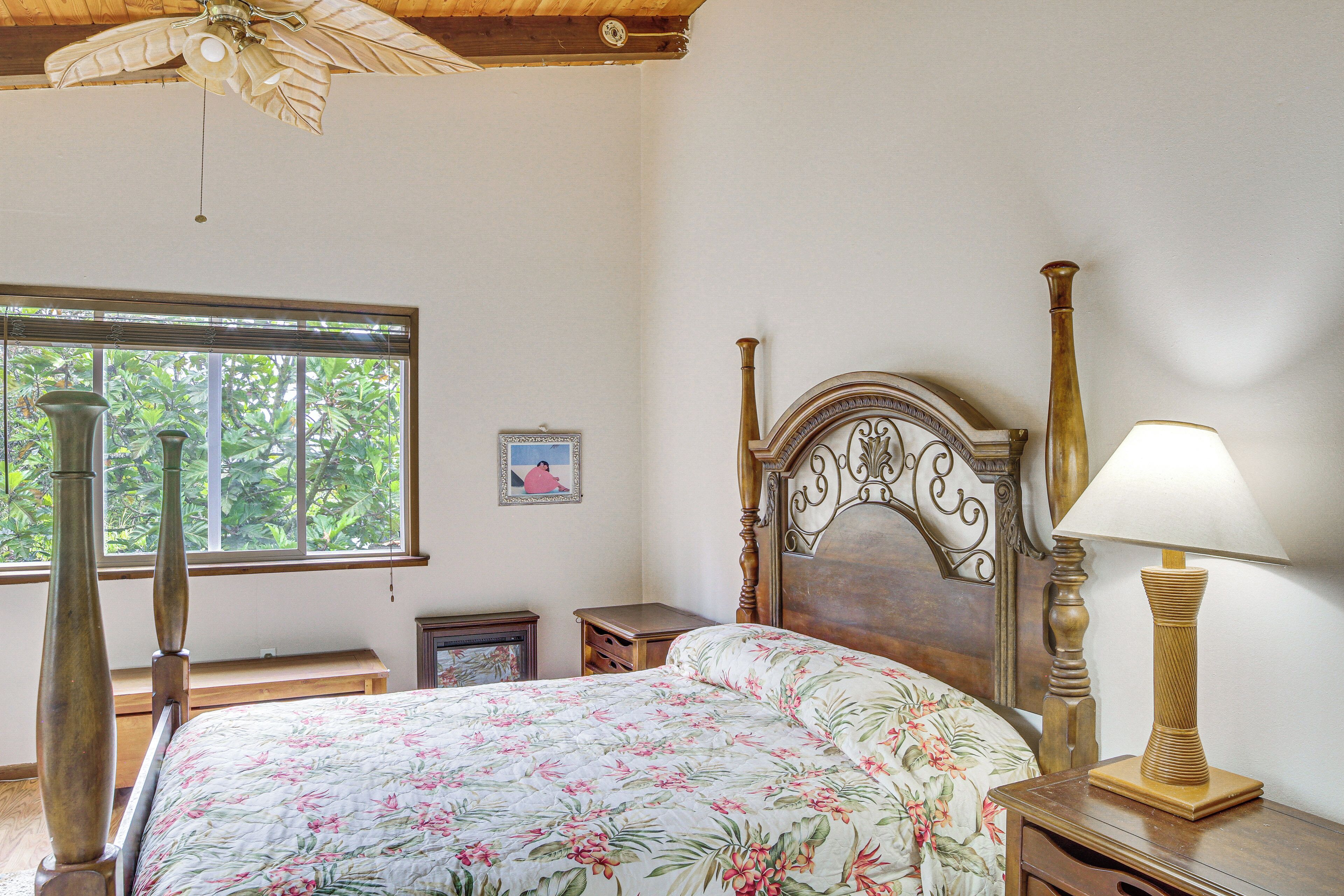 Een slaapkamer met een houten bed, een bedlampje, een nachtkastje, een raam met uitzicht op groen en een plafondventilator.