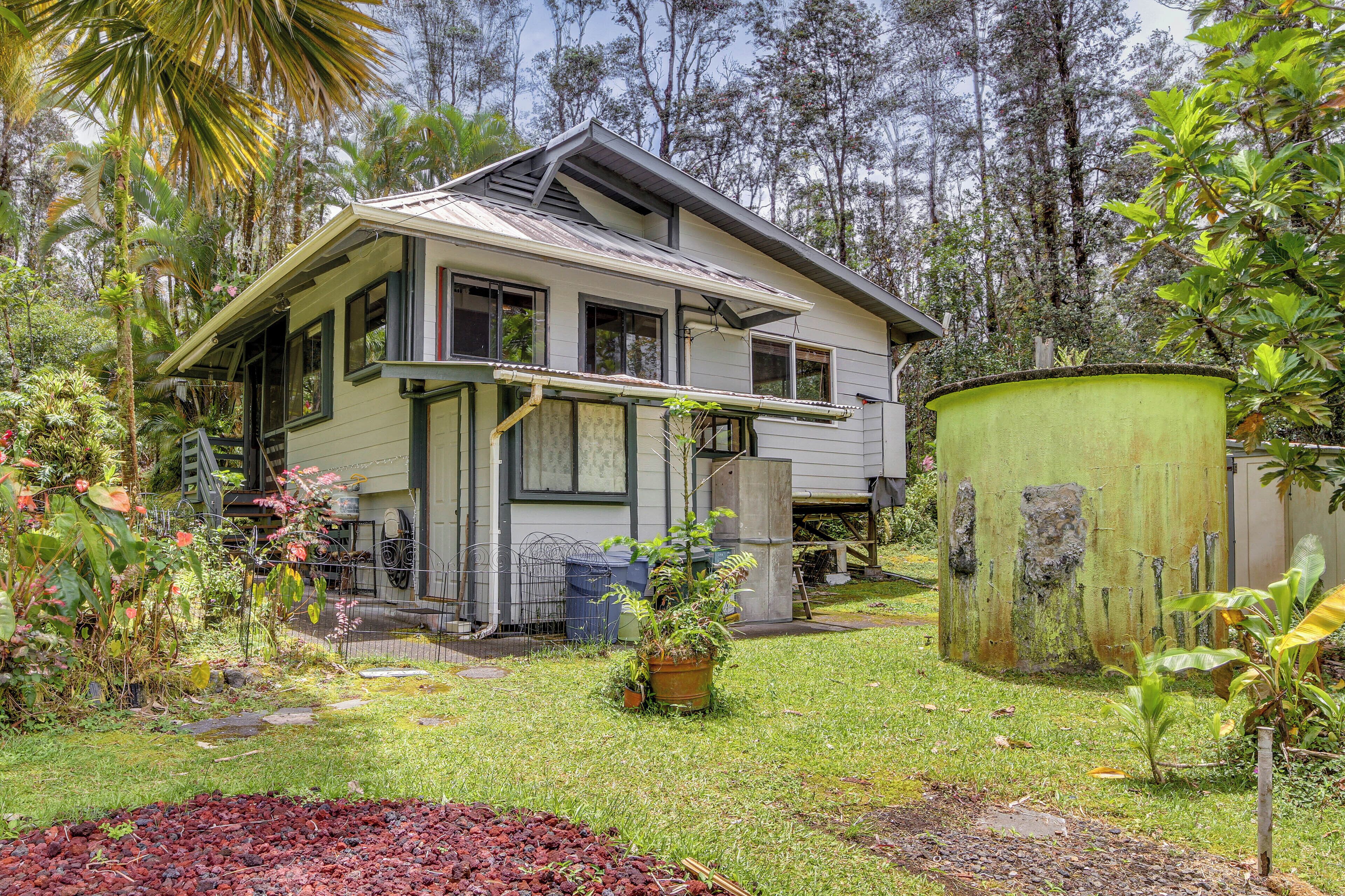 A house with a sloped roof, a porch, and a yard with plants and a green barrel.