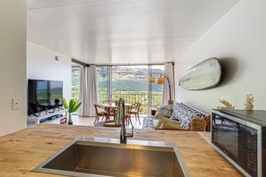 A modern kitchen with a large window, a dining area, and a living room with a view. - Stay on Waianae Coast! Ocean-view Condo w/ Pools (Waianae)