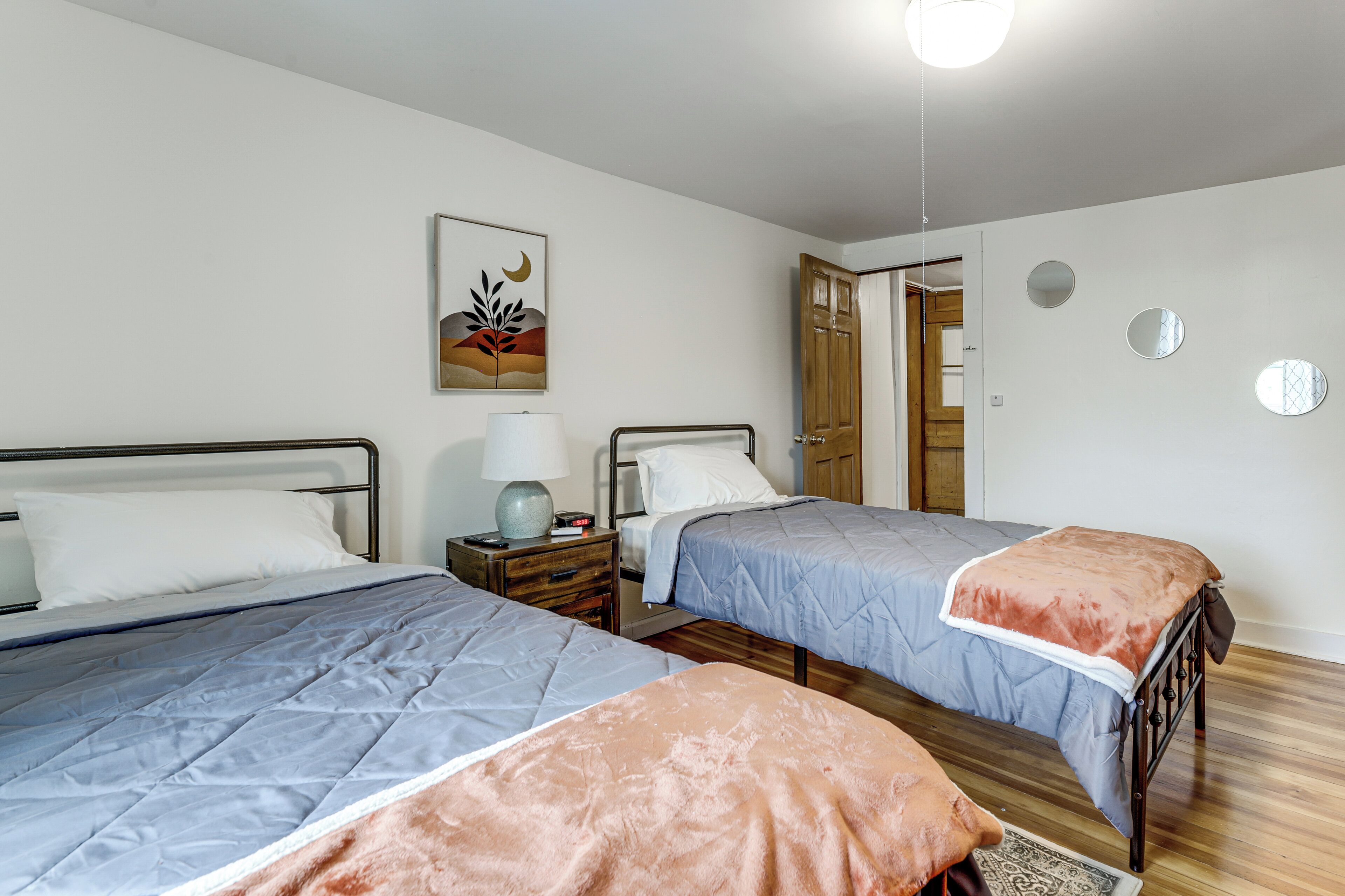 A bedroom with two beds, a nightstand, a lamp, a door, and a wall with a circular mirror and a painting.