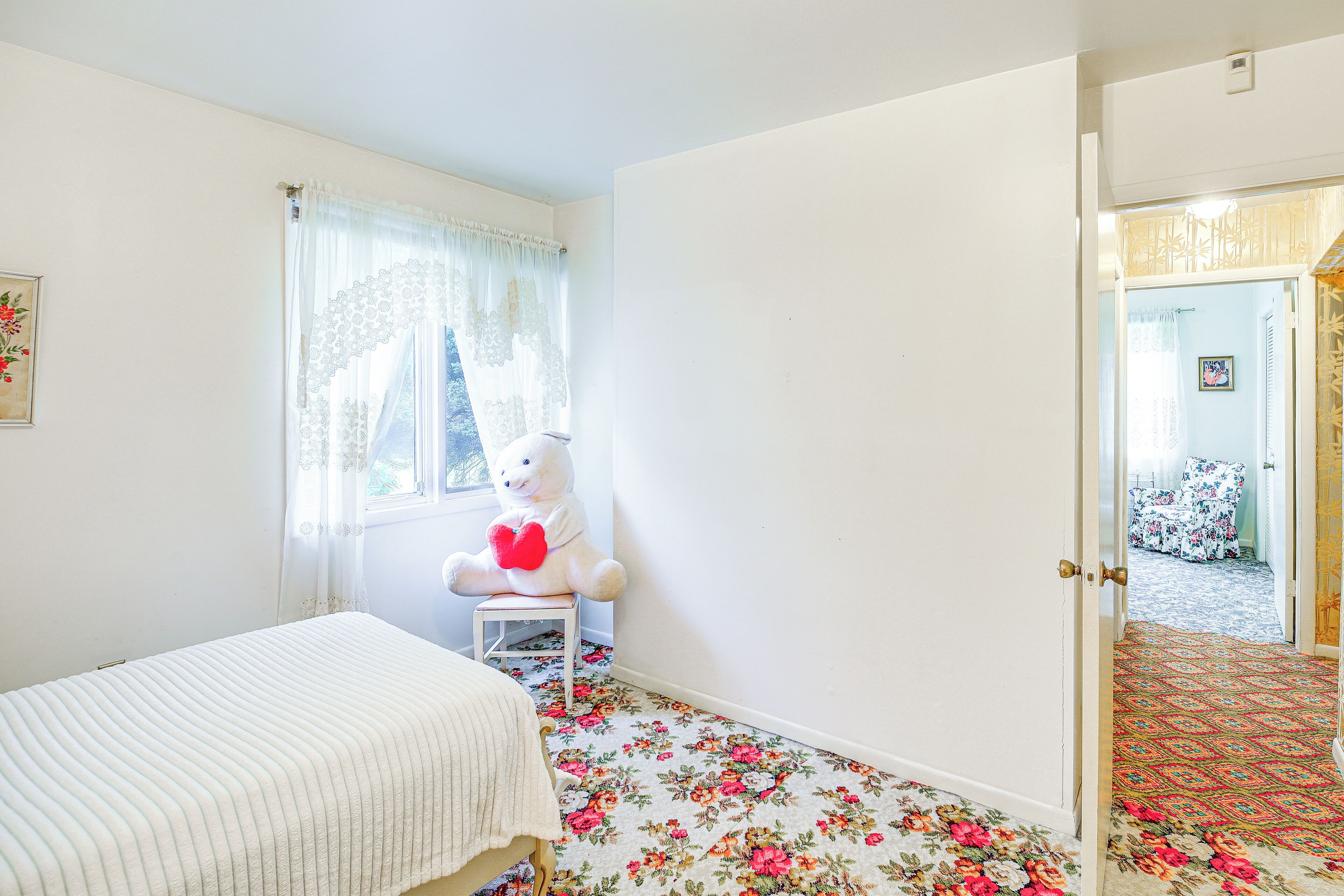 Ein Schlafzimmer mit einem Bett, einem Sessel mit Teddybär, einem Fenster mit Vorhängen und einer Tür zu einem anderen Raum.
