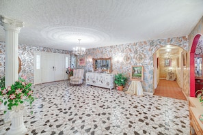 A room with a patterned floor, a white column, a chandelier, a cabinet with artwork, and a mirror.