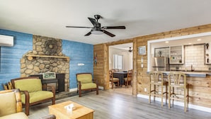 A living room with a stone fireplace, wooden furniture, and a kitchen area with a refrigerator and bar.