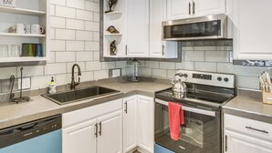 A kitchen with white cabinets, a stainless steel microwave, a kettle, and a dish rack.