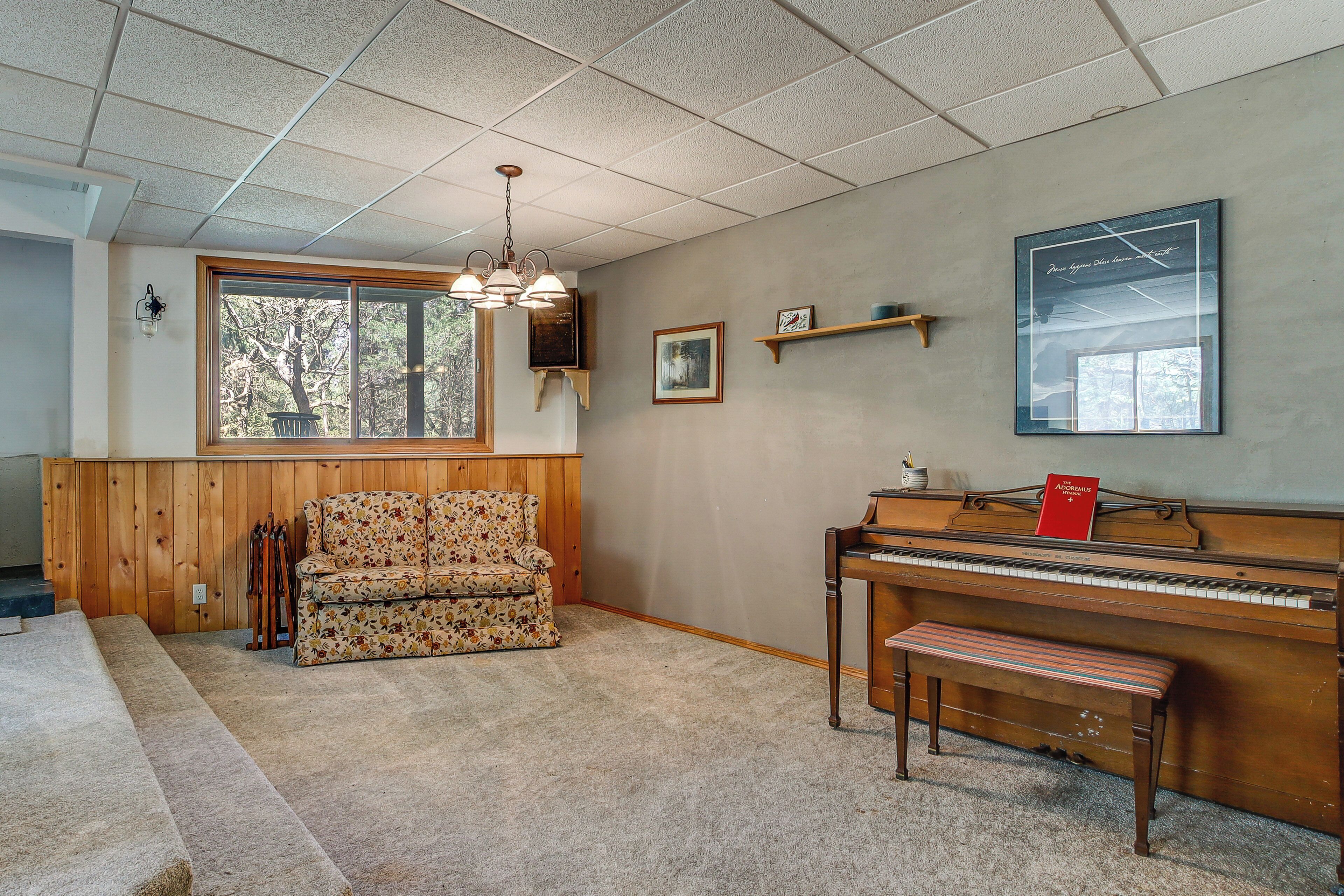 A room with a piano, a bench, a floral sofa, a wooden wall, a window, and a ceiling light.