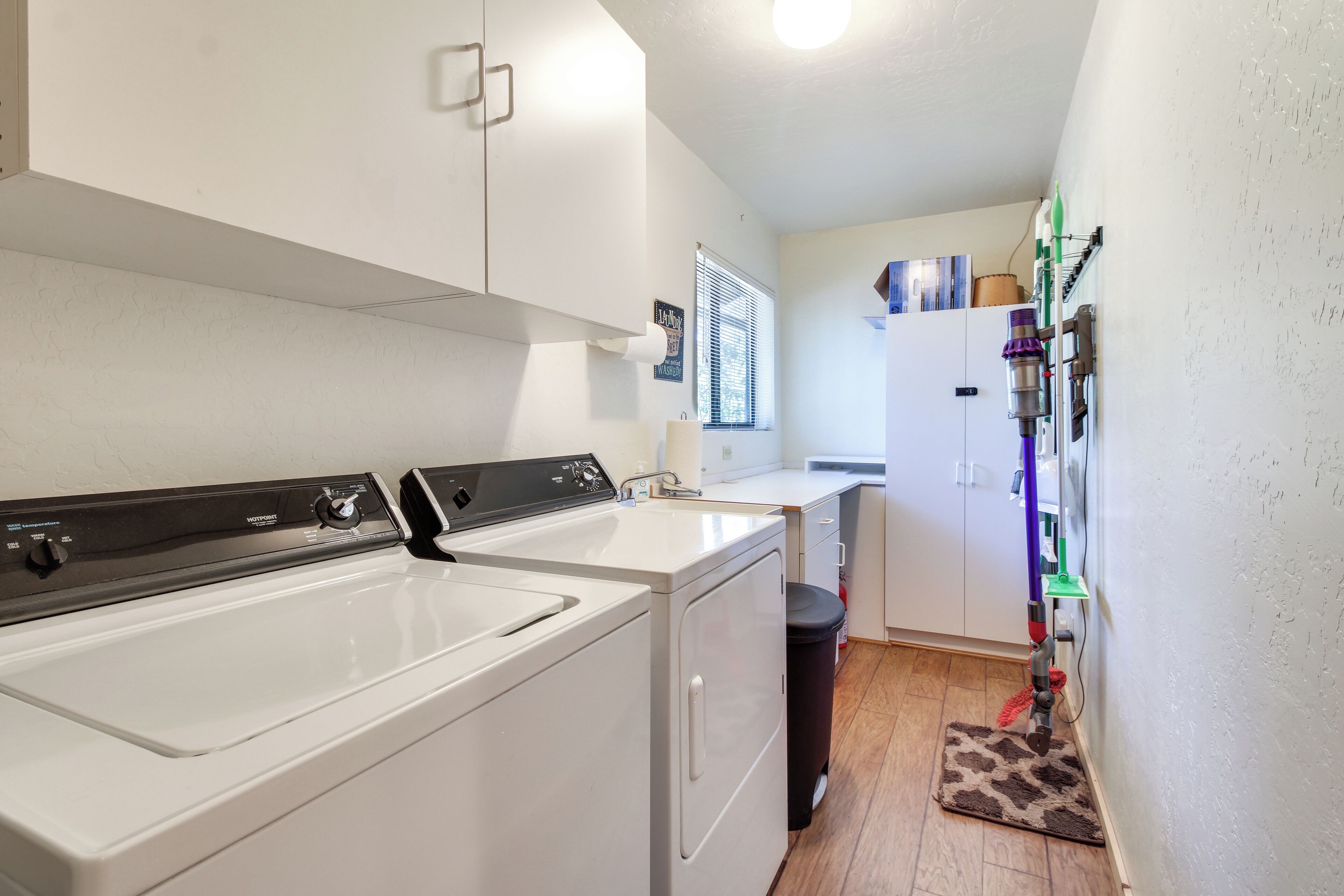 A laundry area with two washers and dryers, a window with blinds, a small rug, and a wall-mounted cleaning tool.