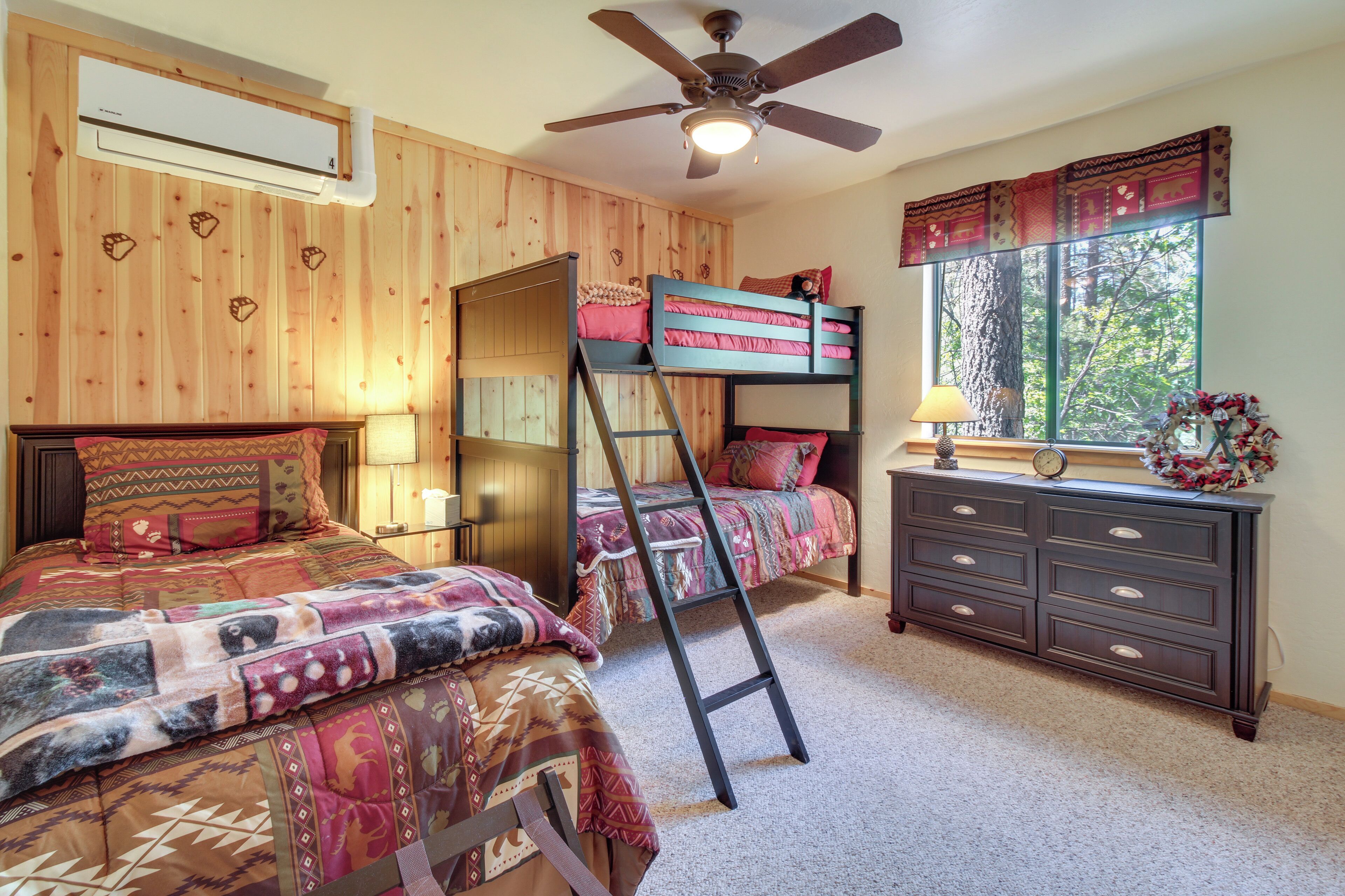 A bedroom with a bunk bed, a dresser, a ladder, a ceiling fan, and a window with a view of trees.