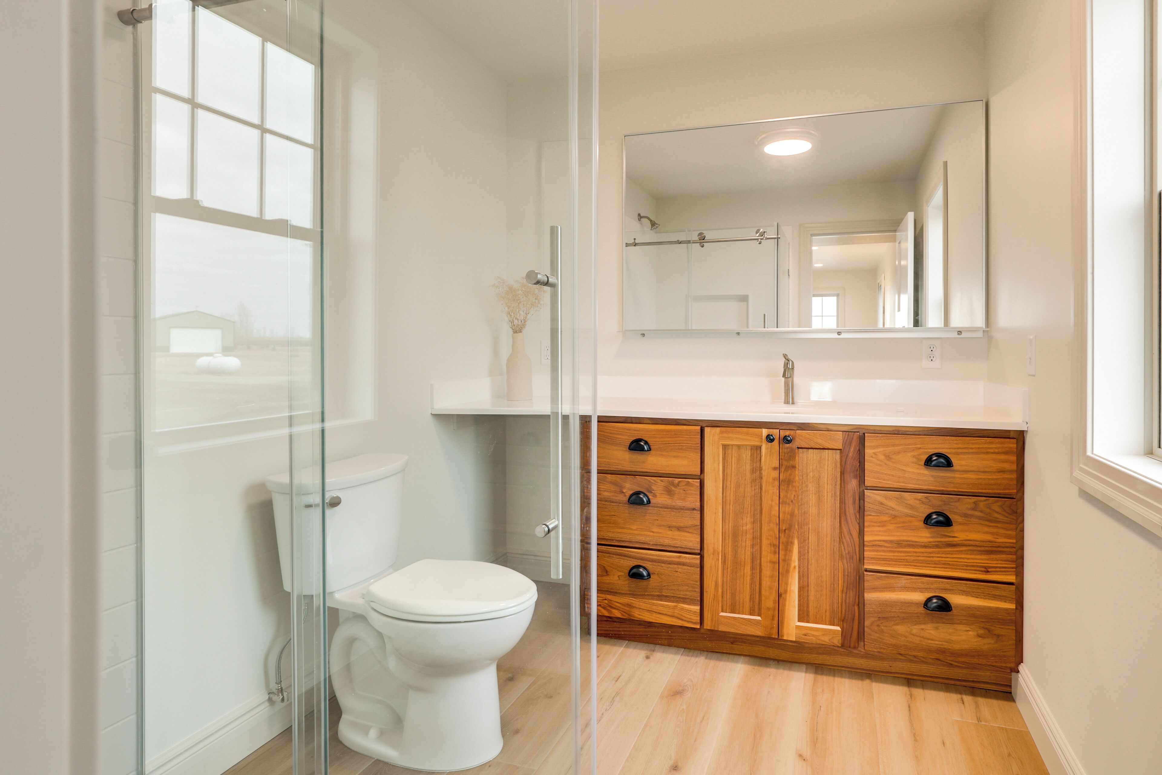 A modern bathroom with a glass shower enclosure, a white toilet, wooden cabinetry, and a large mirror above the sink.